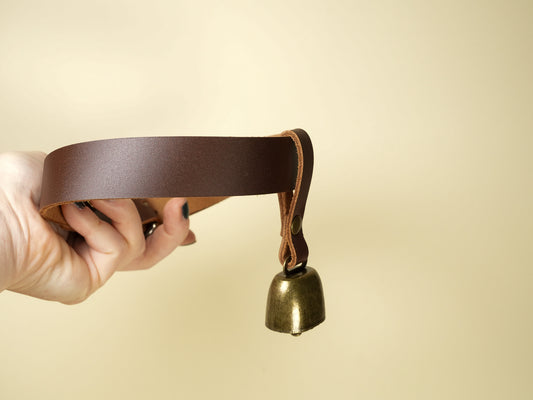 Hand holding a brass cowbell attached to a brown leather strap on beige background