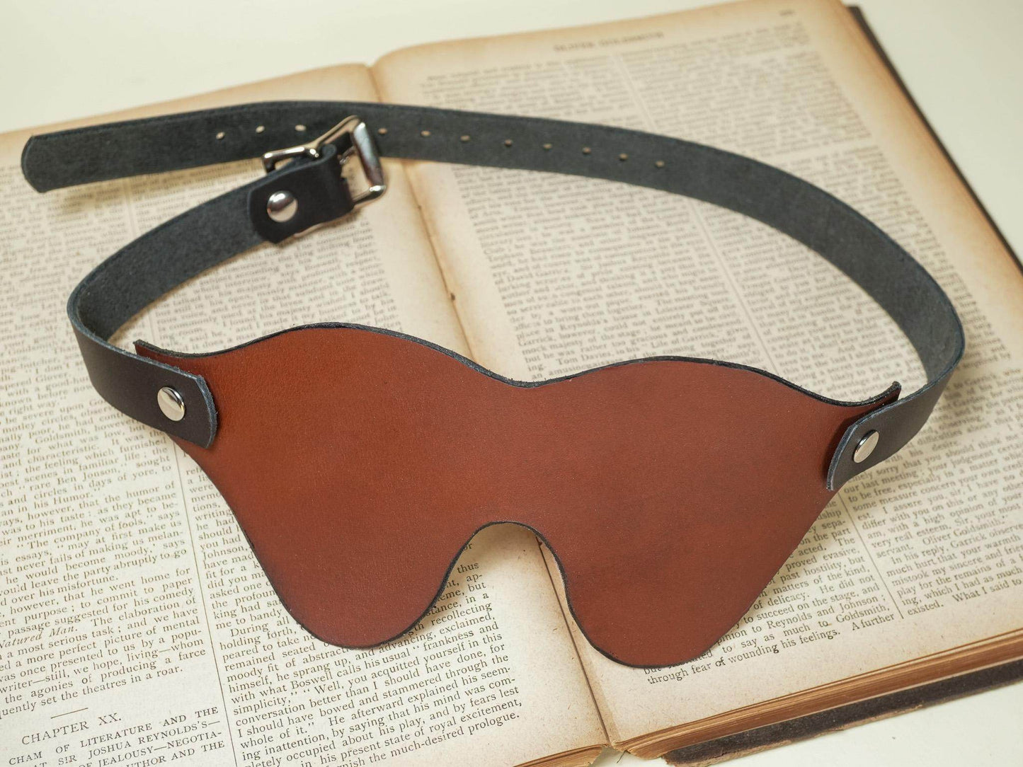 Bound by Dave leather blindfold in brown with adjustable black strap laid on an open book