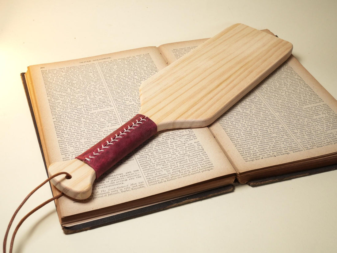 Sadistic wood paddle with red leather-wrapped handle resting on an open vintage book