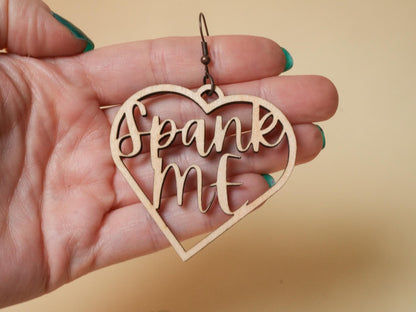 Wooden heart-shaped BDSM earrings with spank me text held in hand by Bound by Dave
