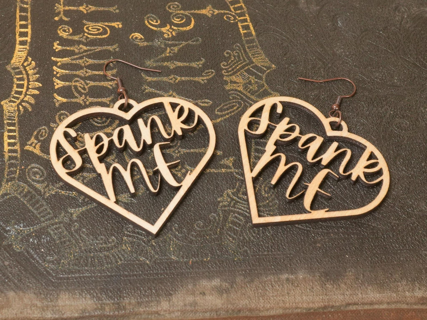 Bound by Dave wooden heart-shaped bdsm earrings with cutout text Spank Me on textured surface