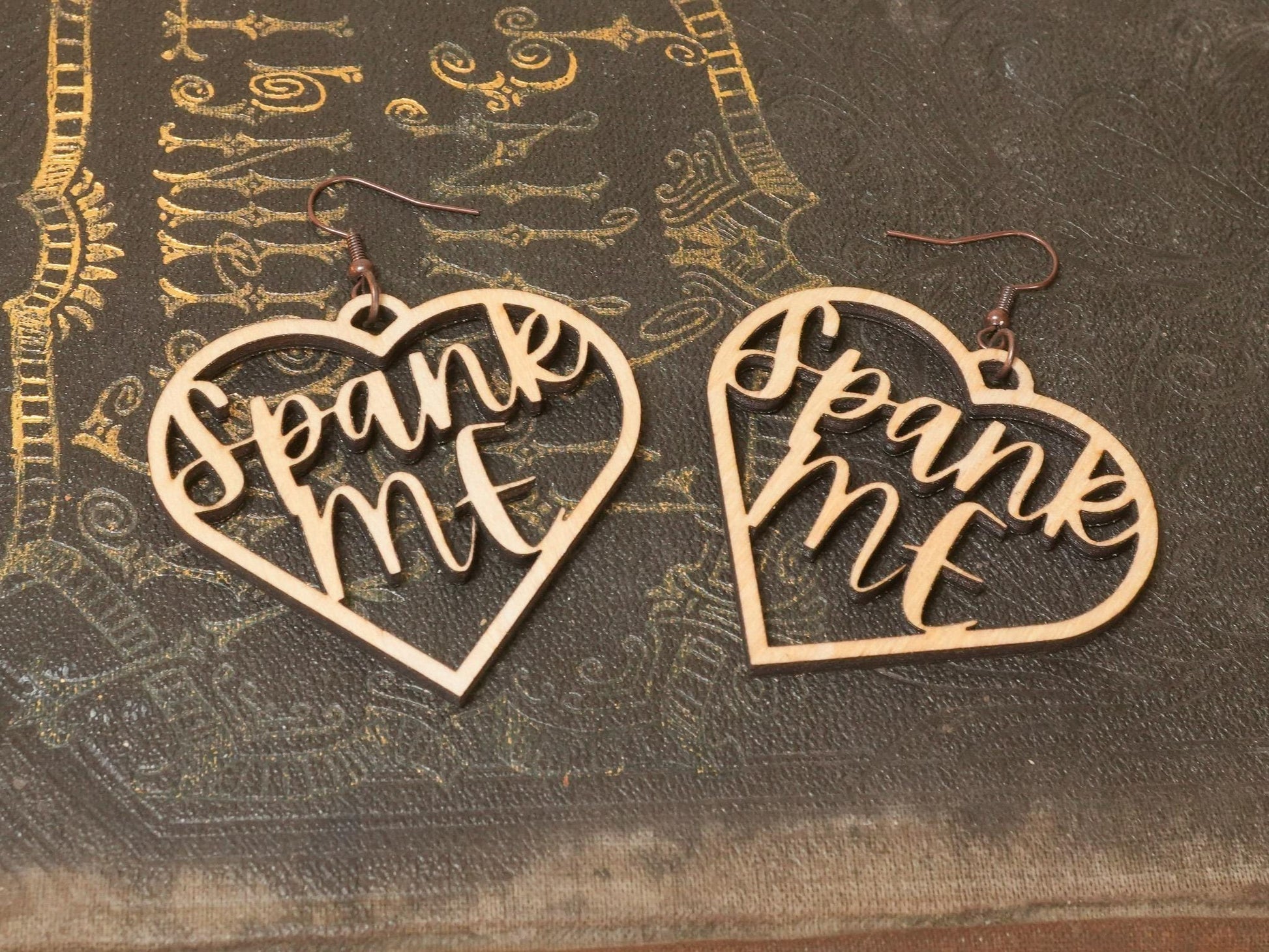 Bound by Dave wooden heart-shaped bdsm earrings with cutout text Spank Me on textured surface