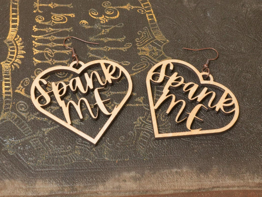 Bound by Dave wooden heart-shaped bdsm earrings with cutout text Spank Me on textured surface