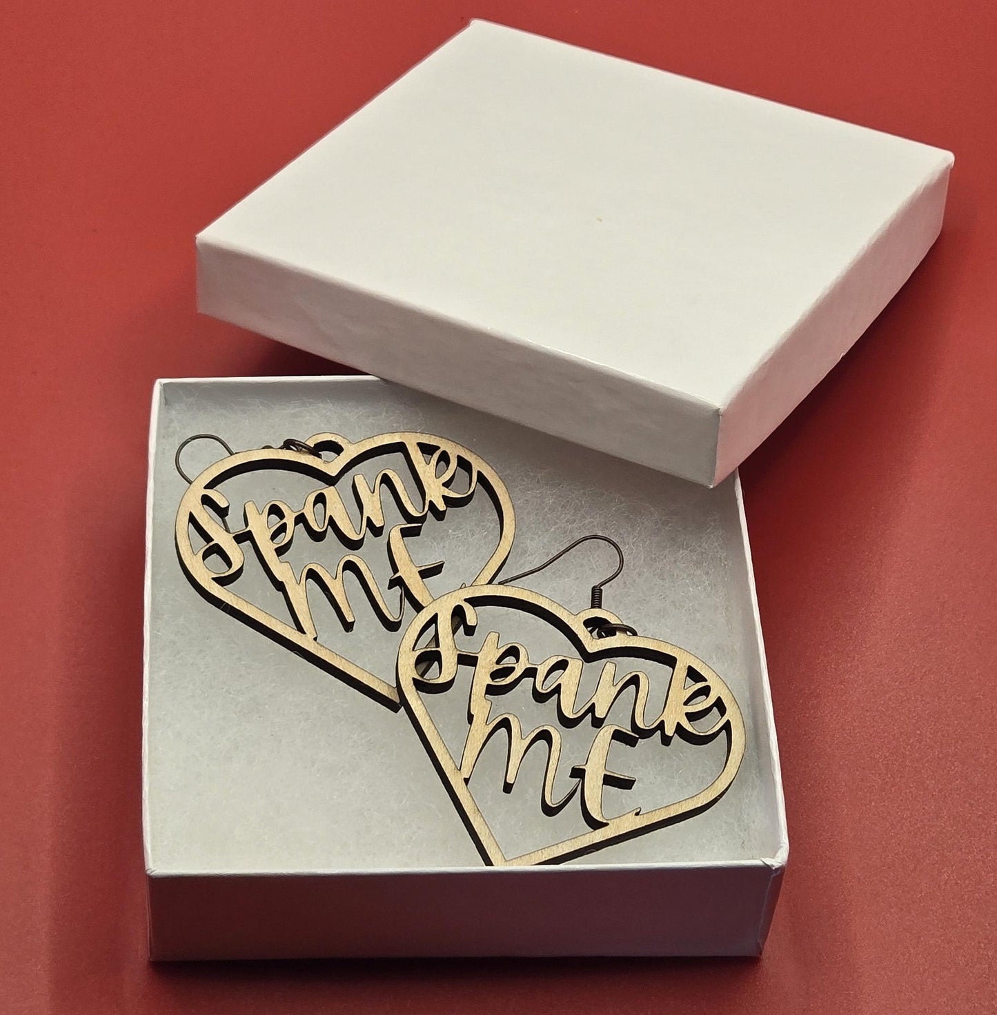 Bound by Dave wooden bdsm earrings in heart shape with Spank Me text inside displayed in white box