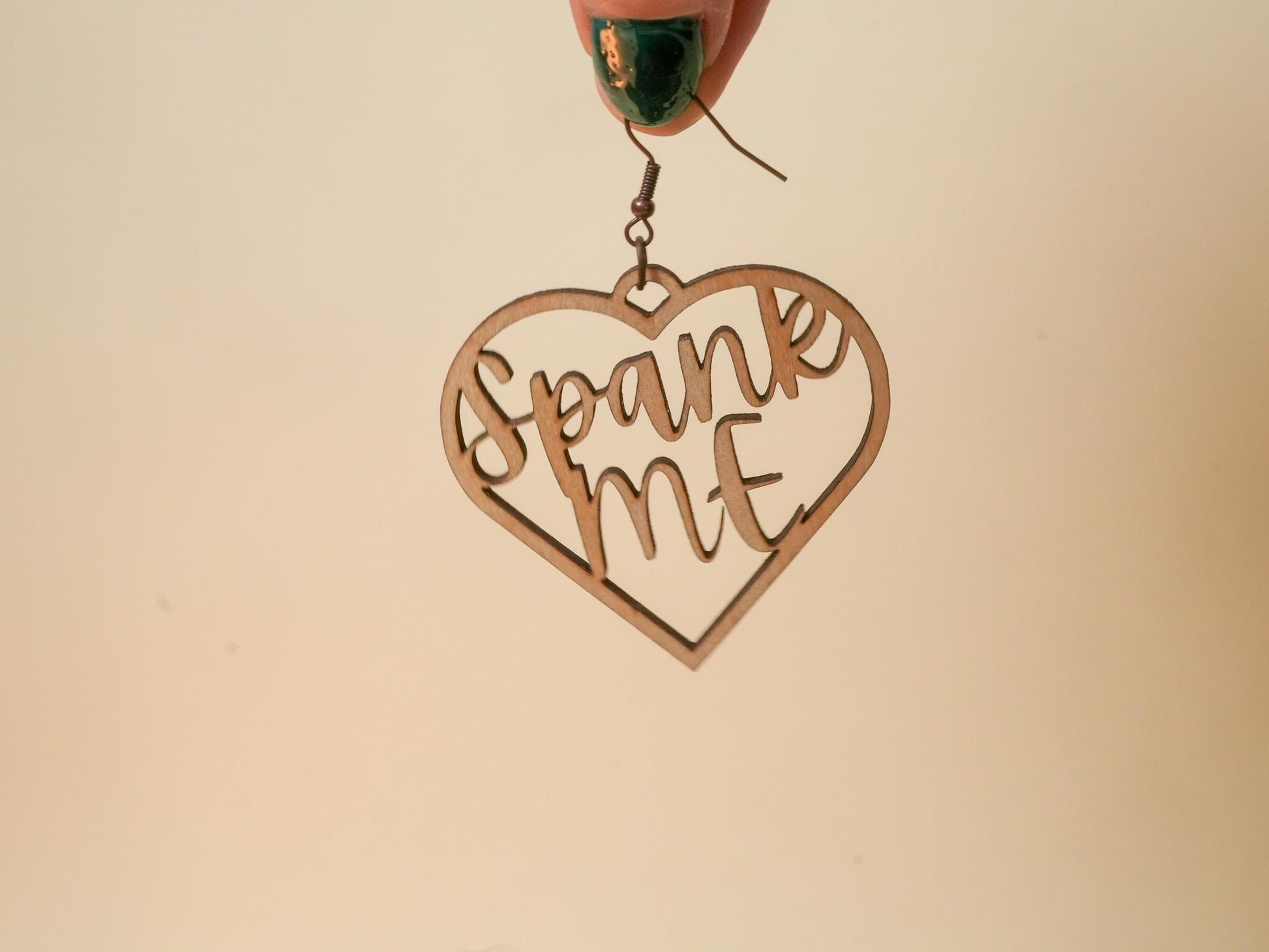 Heart-shaped wooden BDSM earrings with cutout text spank me held by hand with green nail polish