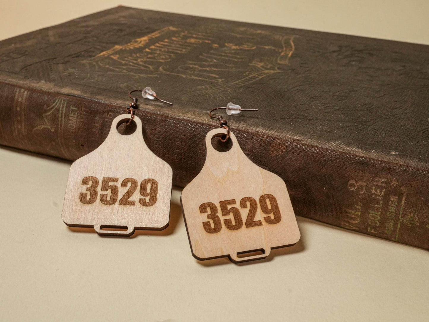 Wooden cow tag earrings with the number 3529 engraved, displayed on a vintage book background