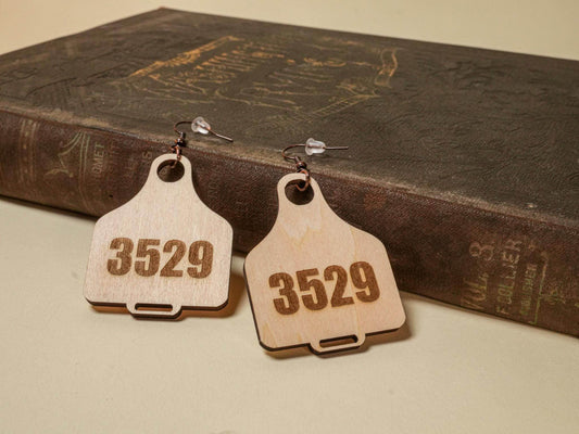 Wooden cow tag earrings with the number 3529 engraved, displayed on a vintage book background