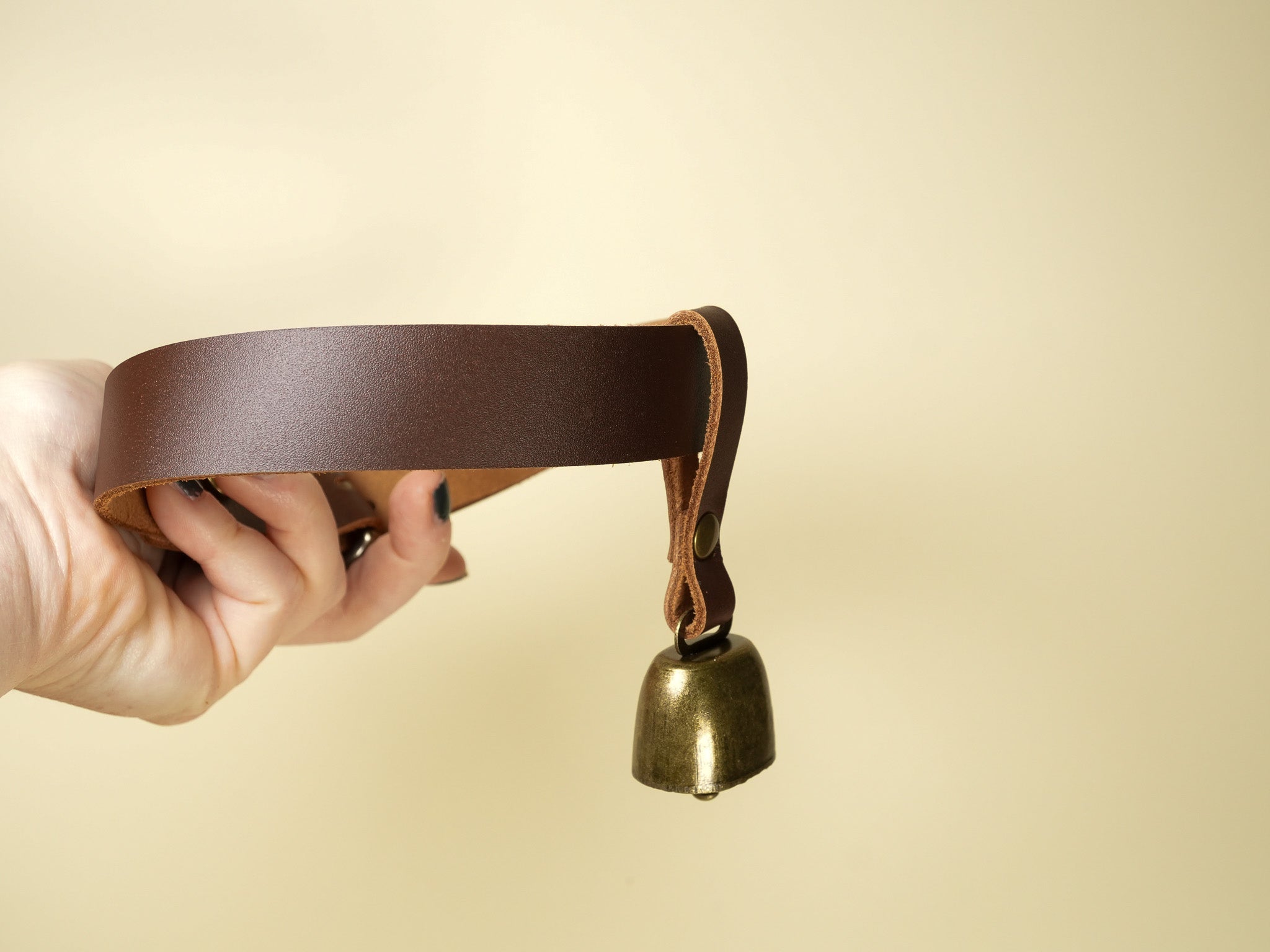 Hand holding a brass cowbell attached to a brown leather strap on beige background