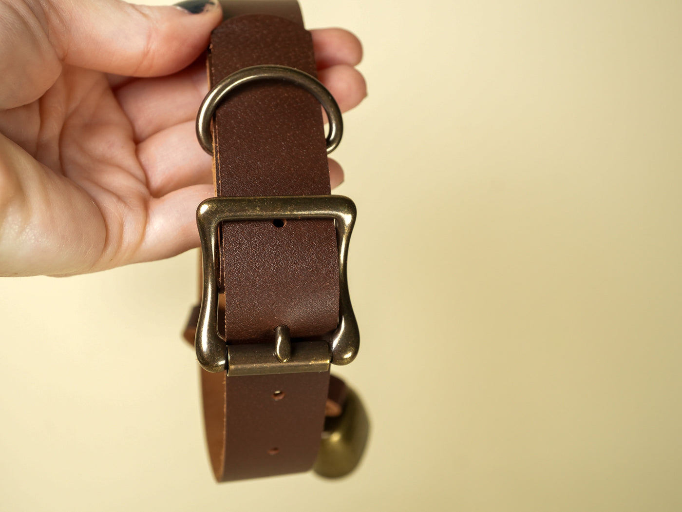 Brown leather dog collar with brass buckle and attached cowbell held in hand by Bound by Dave