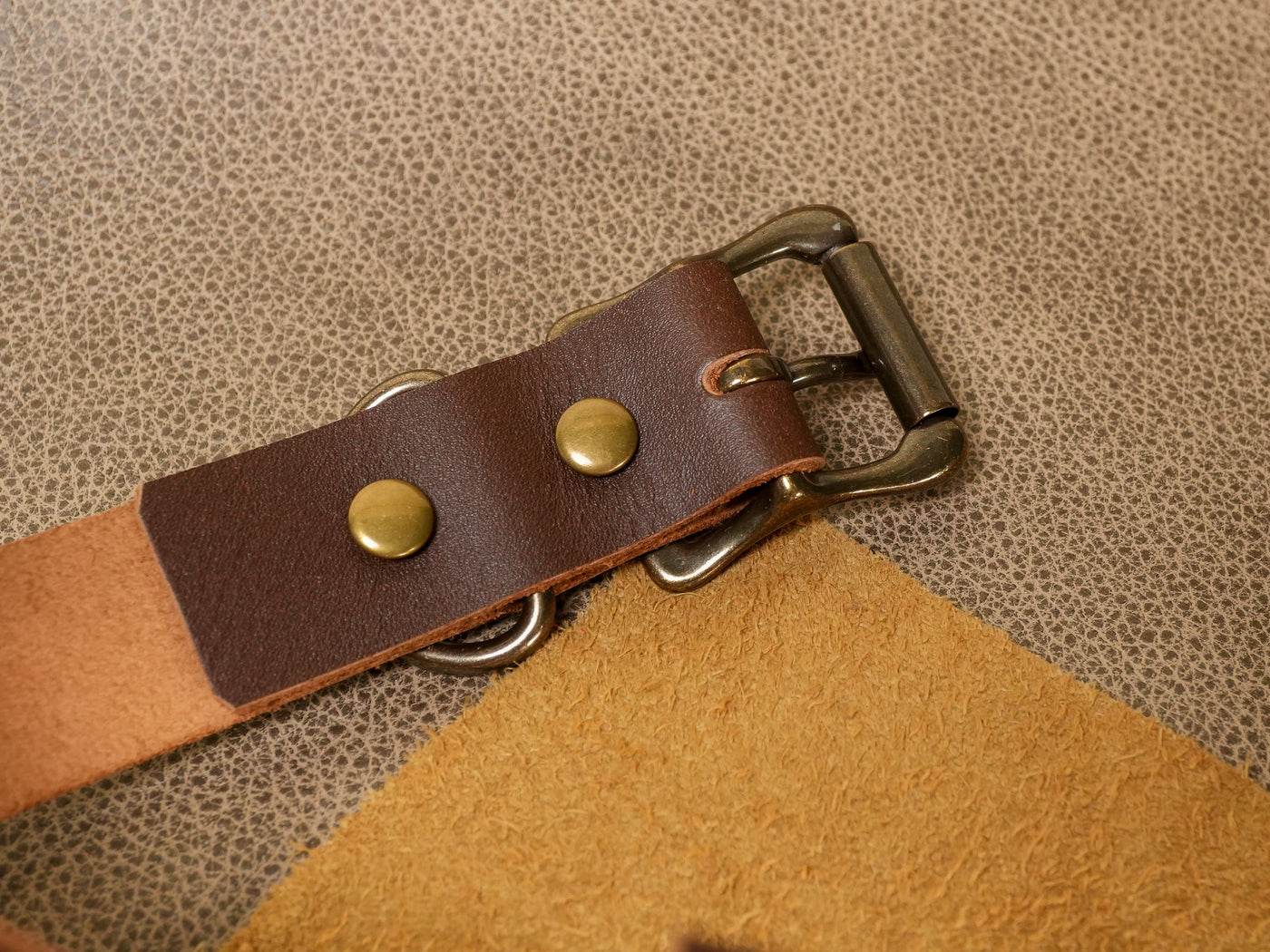Close-up of Bound by Dave leather cowbell strap with brass rivets and metal buckle on textured surface