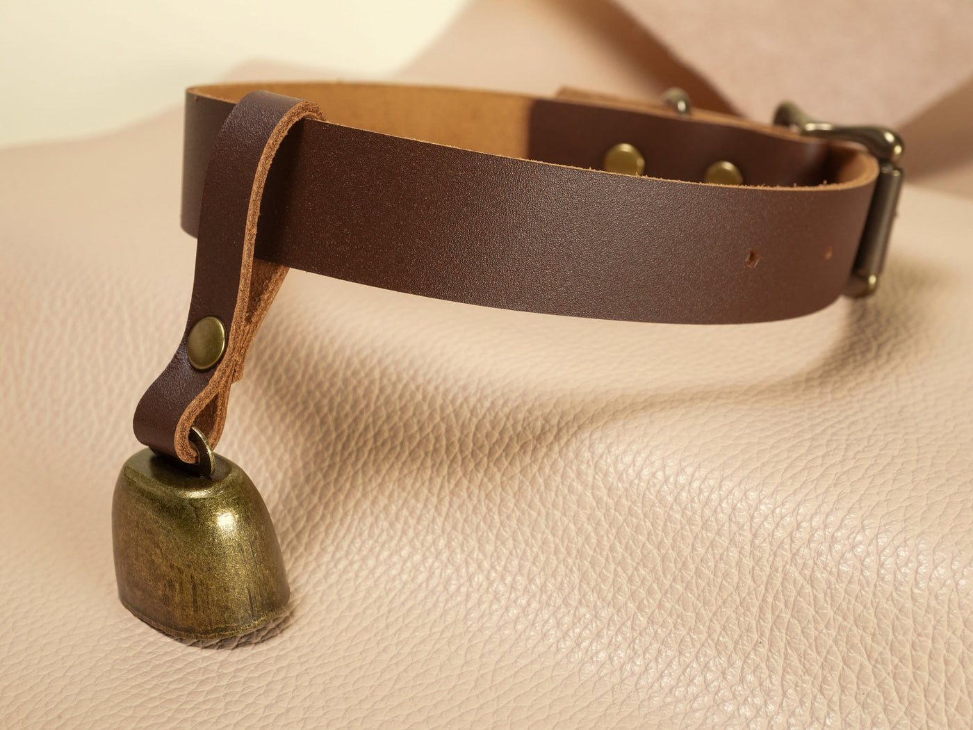 Bound by Dave brown leather strap collar with attached metal cowbell on beige textured surface