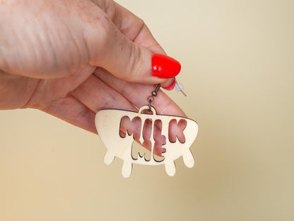 Hand holding Bound by Dave Hucow Earrings shaped like a cow udder with milk me cutout