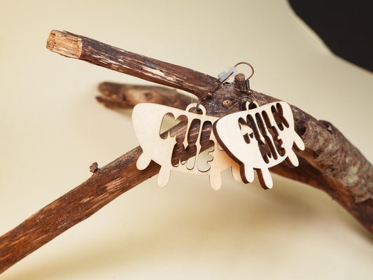 Hucow Earrings by Bound by Dave wooden earrings with milk me cutout design hanging on a rustic branch