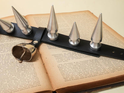 Bound by Dave hucow leather strap with large metal spikes and attached metal bell on open book