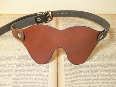Brown Leather Blindfold by Bound by Dave with adjustable buckle strap on an open book background