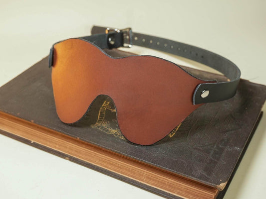 Leather Blindfold by Bound by Dave with adjustable strap resting on a closed vintage book