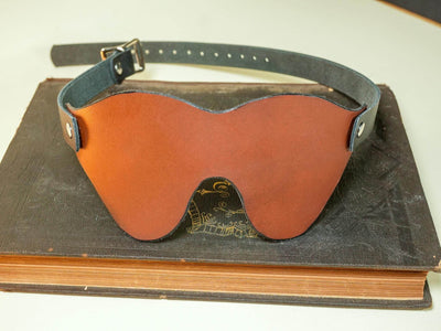 Leather Blindfold by Bound by Dave in brown with adjustable black strap resting on a closed vintage book