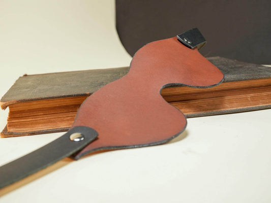 Leather Blindfold in brown and black from Bound by Dave resting on an old book with curved design
