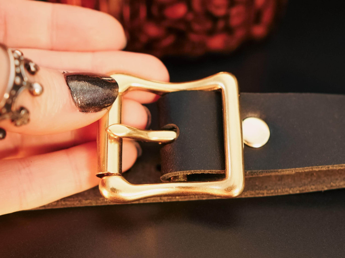 Close-up of a gold buckle on black leather hand cuffs held by a hand with black nail polish