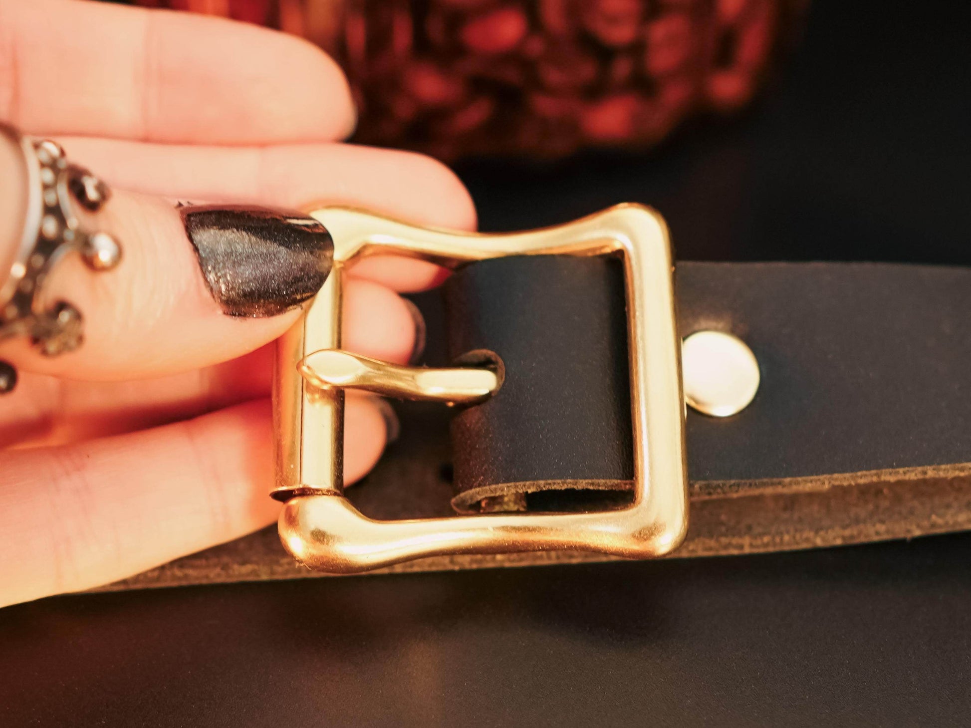 Close-up of a gold buckle on black leather hand cuffs held by a hand with black nail polish