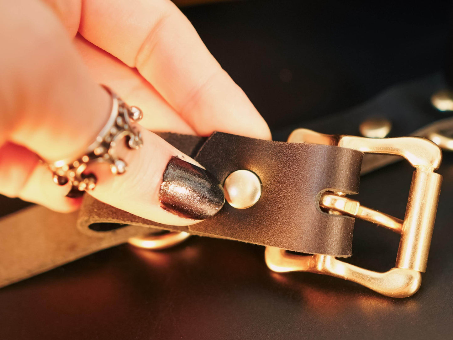 Close-up of a hand adjusting the buckle on black Leather Hand Cuffs with metal rivets and gold hardware