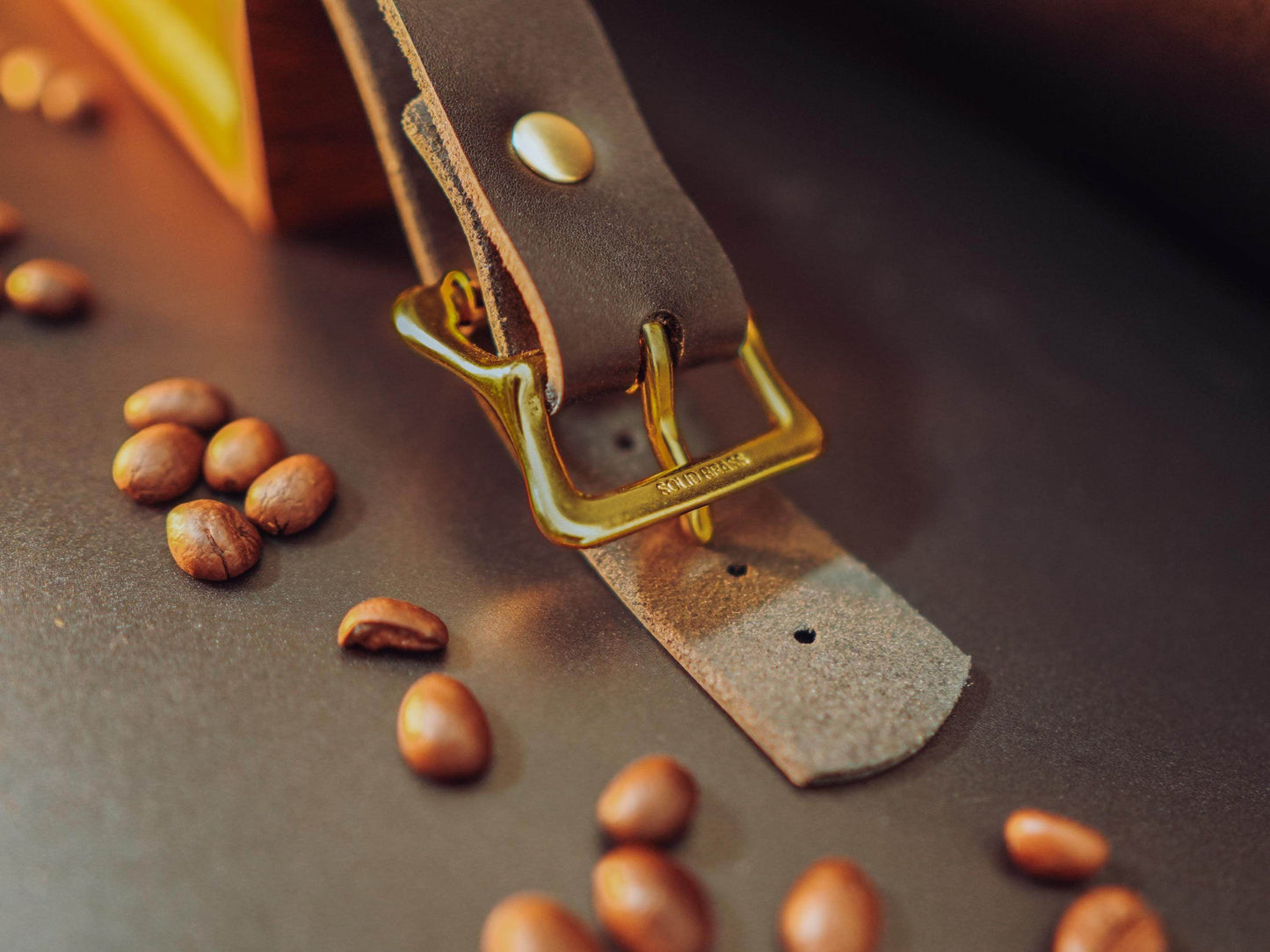 Close-up of brown leather hand cuffs with brass buckle and rivet by Bound by Dave with coffee beans scattered around