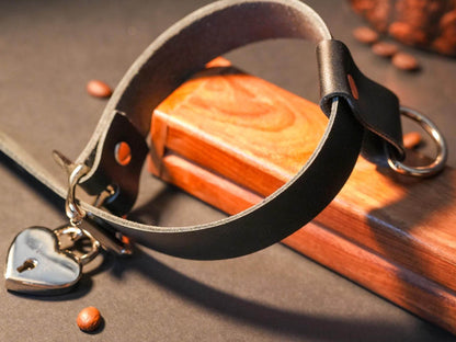 Black leather Locking Sub Collar by Bound by Dave with heart-shaped lock and metal ring on wooden surface