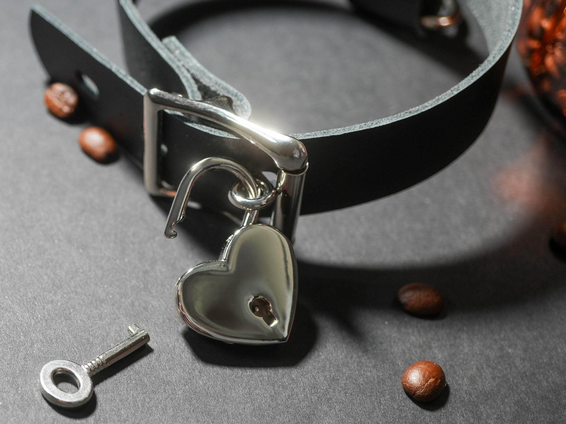 Black leather Locking Sub Collar with heart-shaped silver padlock and key on dark surface with coffee beans