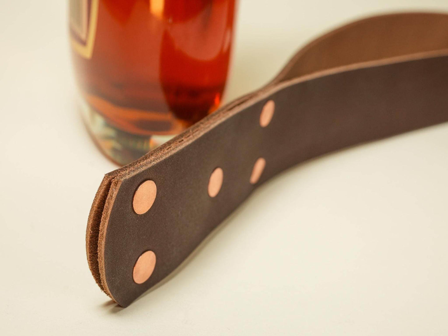 Brown leather spanking belt with copper rivets lying beside a glass bottle