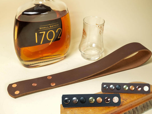 Bound by Dave leather spanking belt with metal studs displayed next to a whiskey bottle and glass