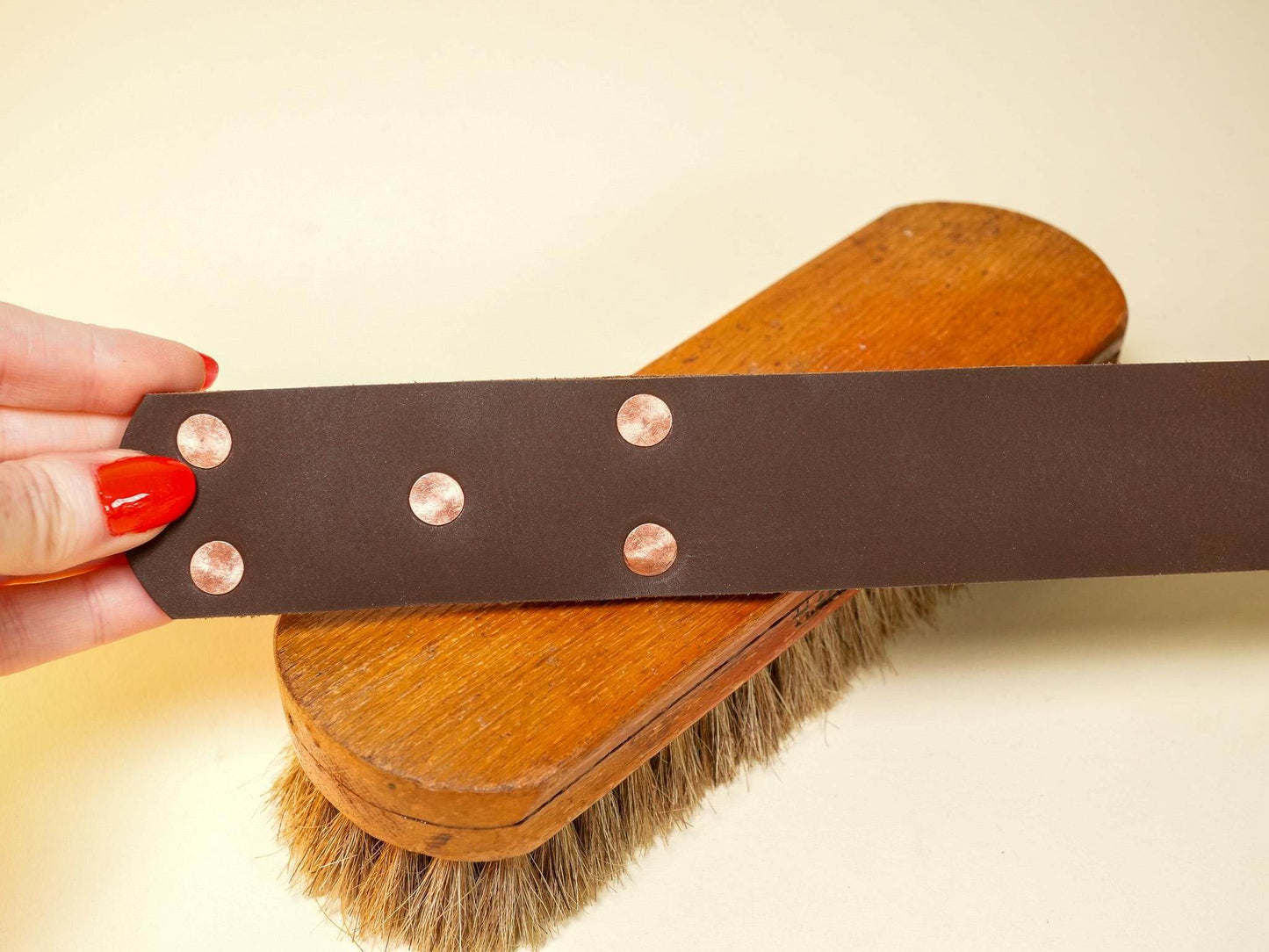 Brown leather spanking belt with copper rivets held above a wooden brush with natural bristles
