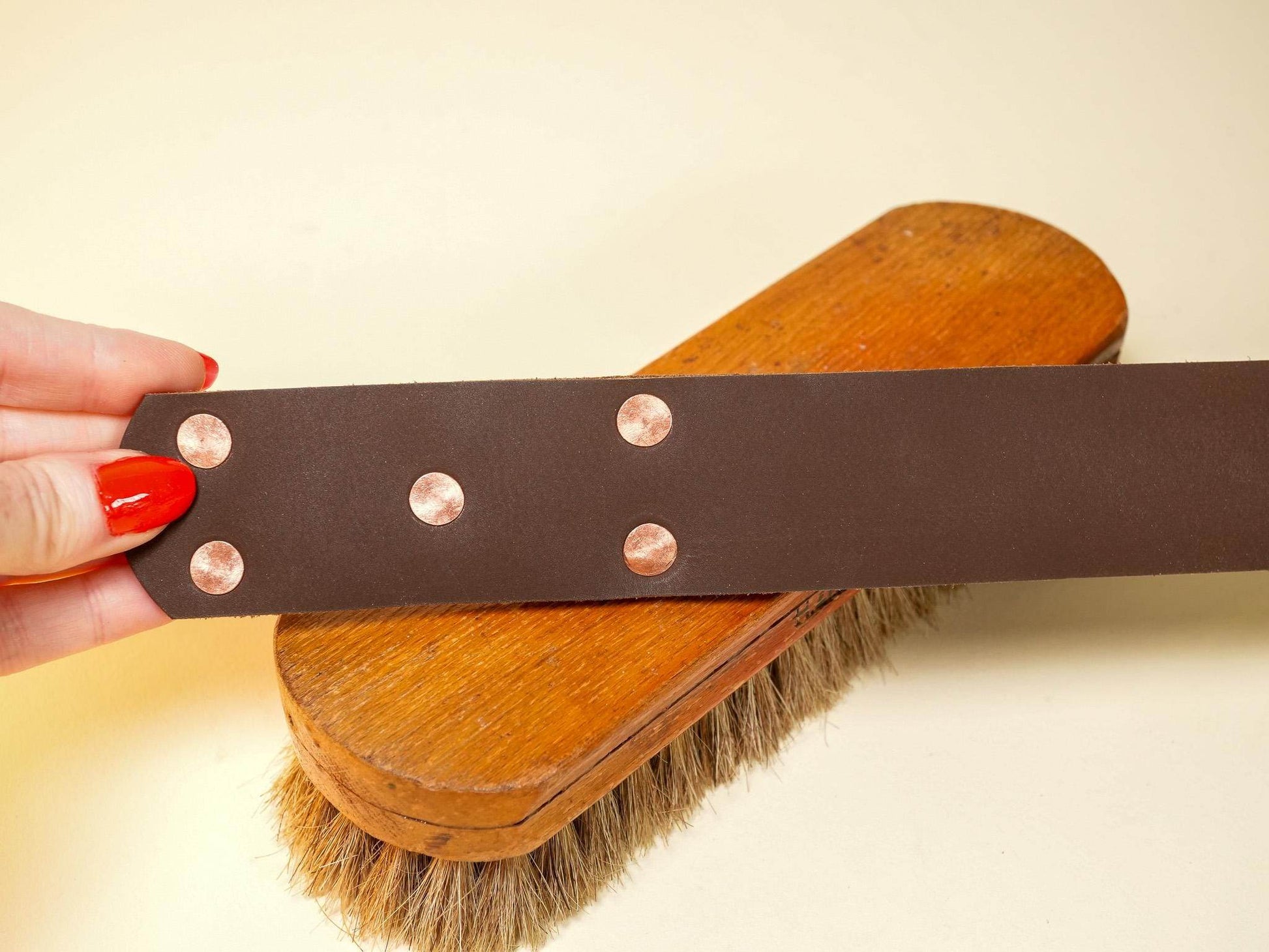 Brown leather spanking belt with copper rivets held above a wooden brush with natural bristles