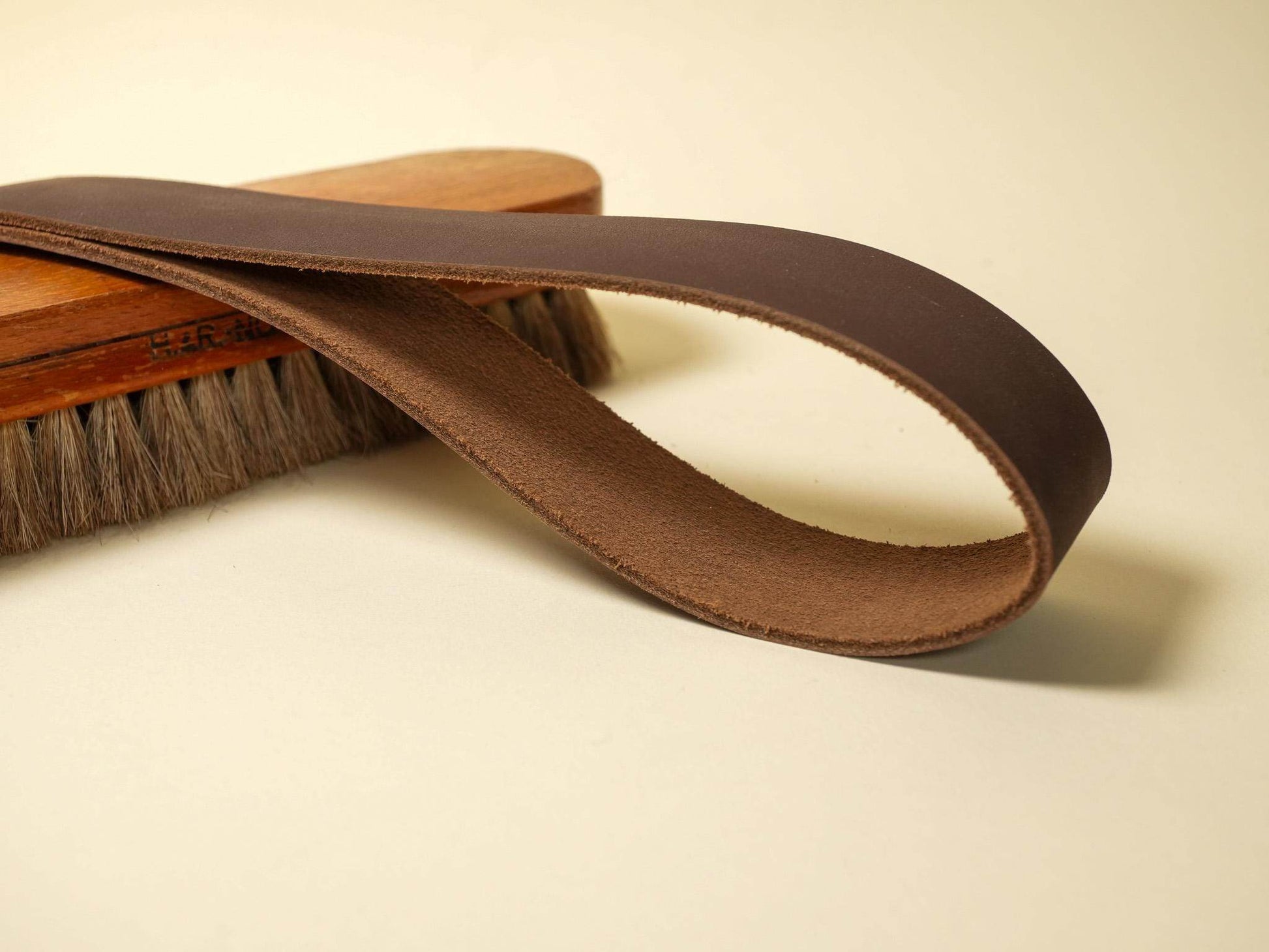 Brown leather spanking belt by Bound by Dave displayed on a wooden brush on light background