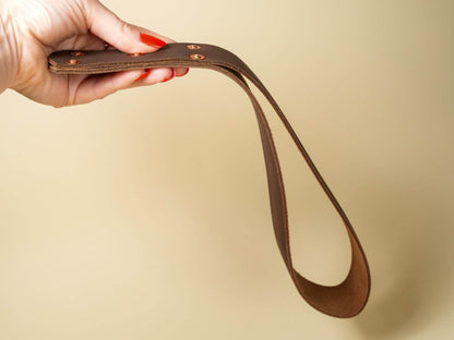 Hand holding a brown leather spanking belt with copper rivets against a beige background
