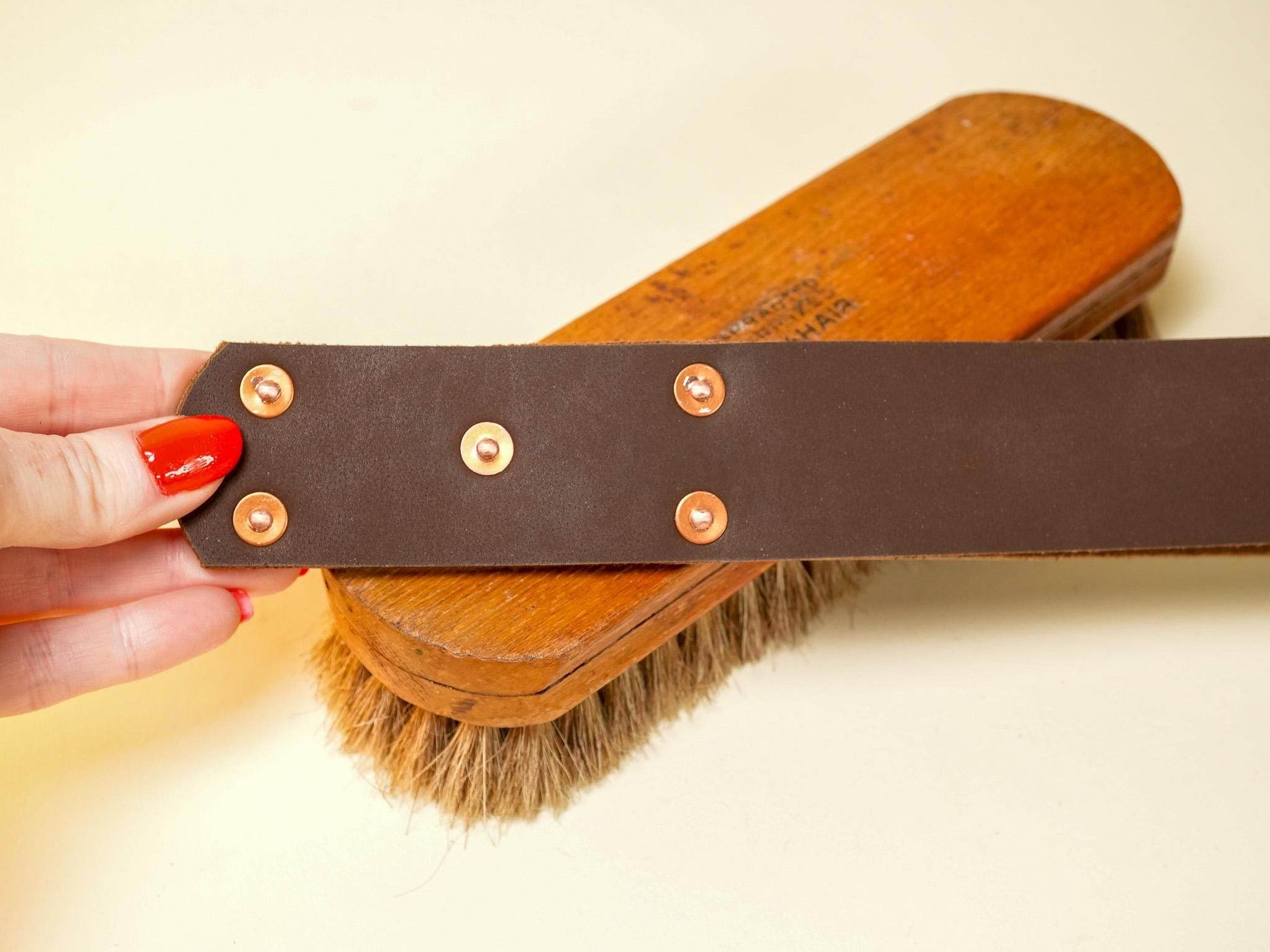 Brown leather spanking belt with brass rivets held above a wooden brush with natural bristles