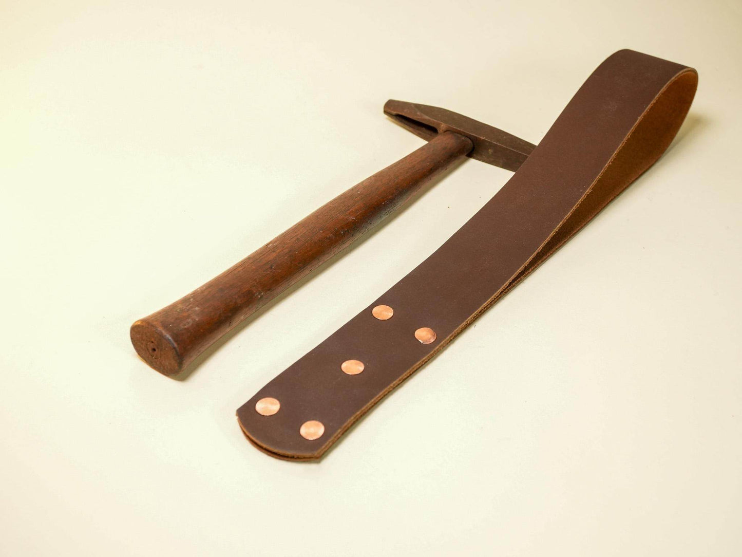 Brown leather spanking belt with copper rivets lying next to a wooden handled hammer on a pale background