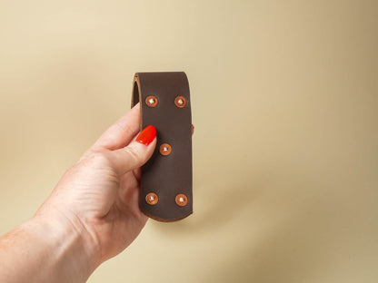Hand holding a brown leather spanking belt with copper rivets on a beige background