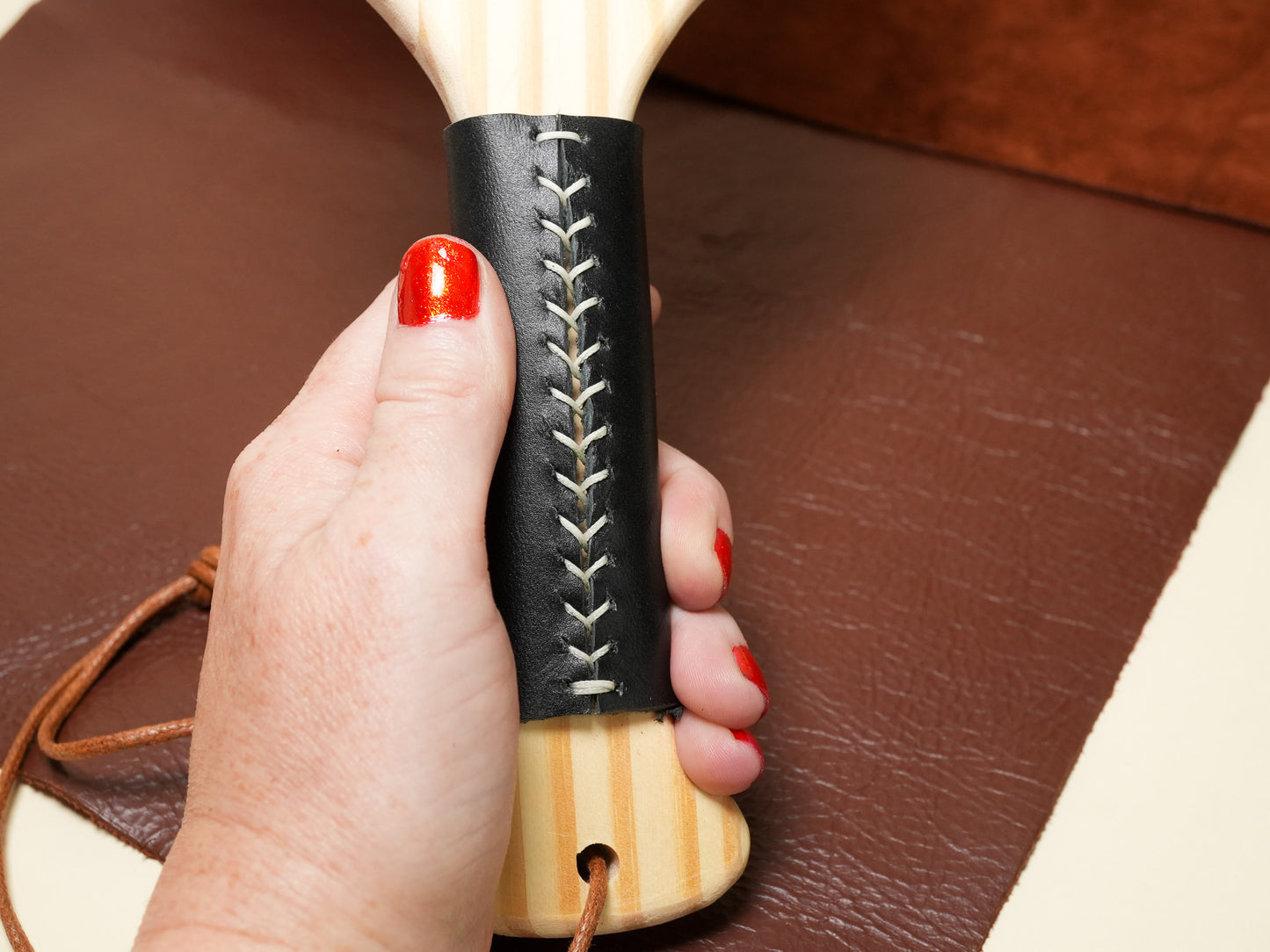 Hand holding a wooden Spanking Paddle with black stitched leather grip on brown background