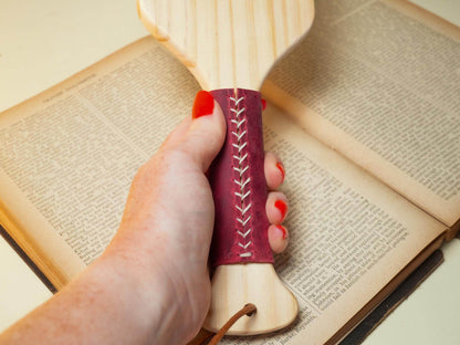 Hand holding wooden spanking paddle with red stitched handle over an open book