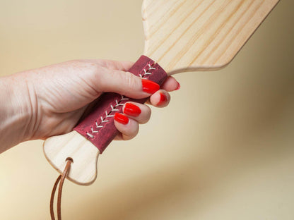 Hand holding a wooden spanking paddle with a red stitched leather grip by Bound by Dave