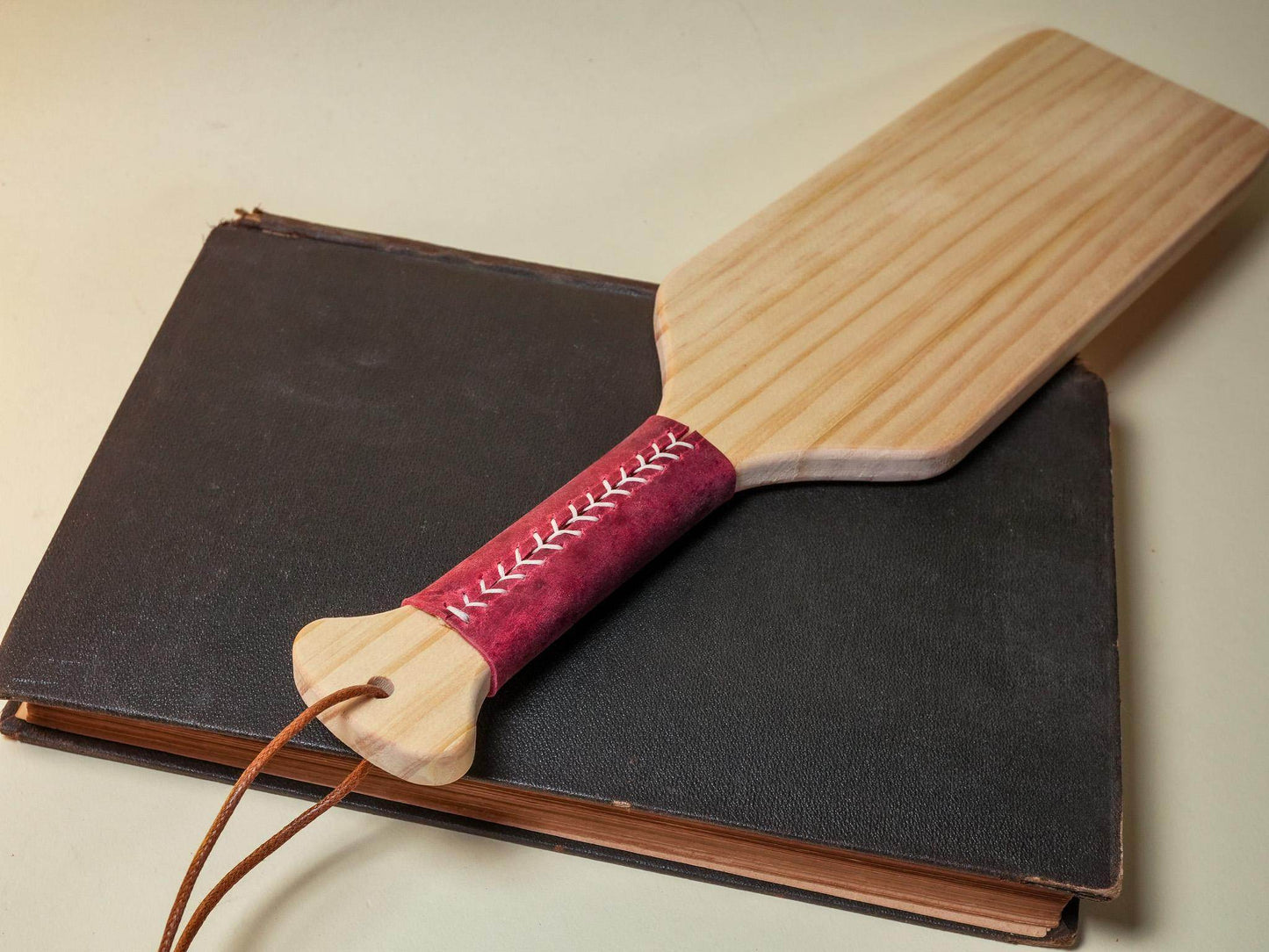 Wooden spanking paddle with red stitched handle by Bound by Dave resting on a closed black book