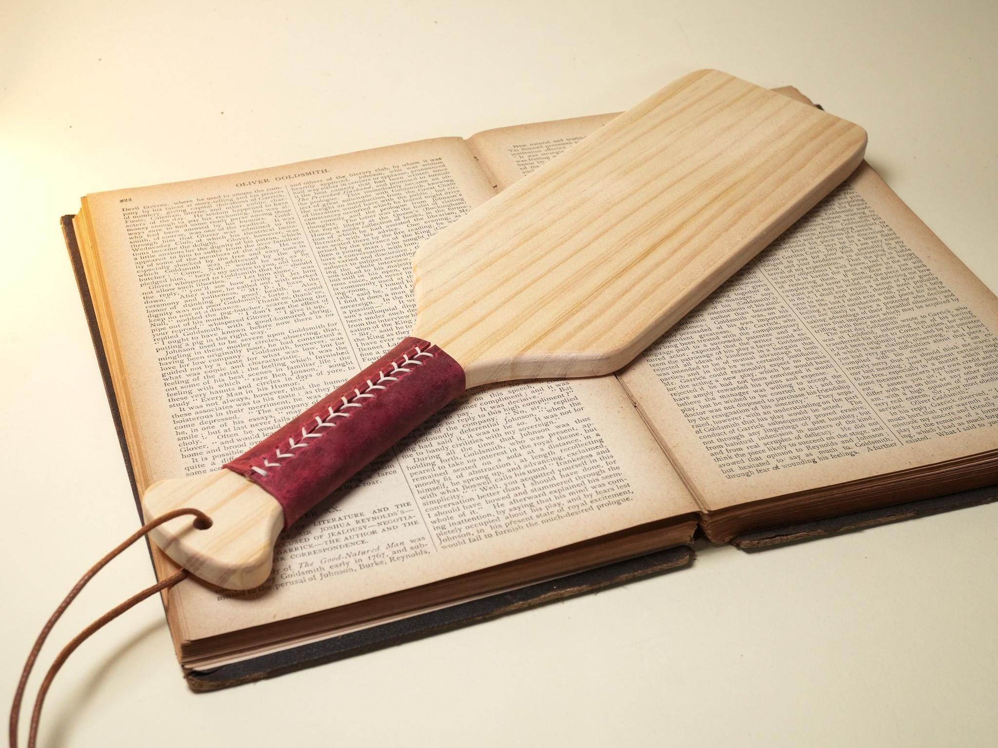 Wooden Spanking Paddle with red stitched leather handle placed on an open book