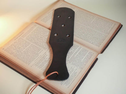 Bound by Dave black leather paddle with six Leather Paddle Holes resting on an open book