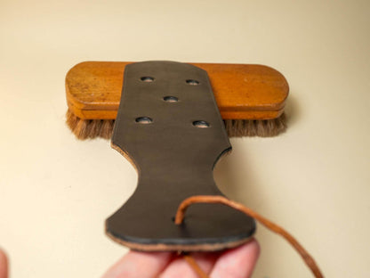 Bound by Dave leather paddle with multiple Leather Paddle Holes and a wooden brush on tan background