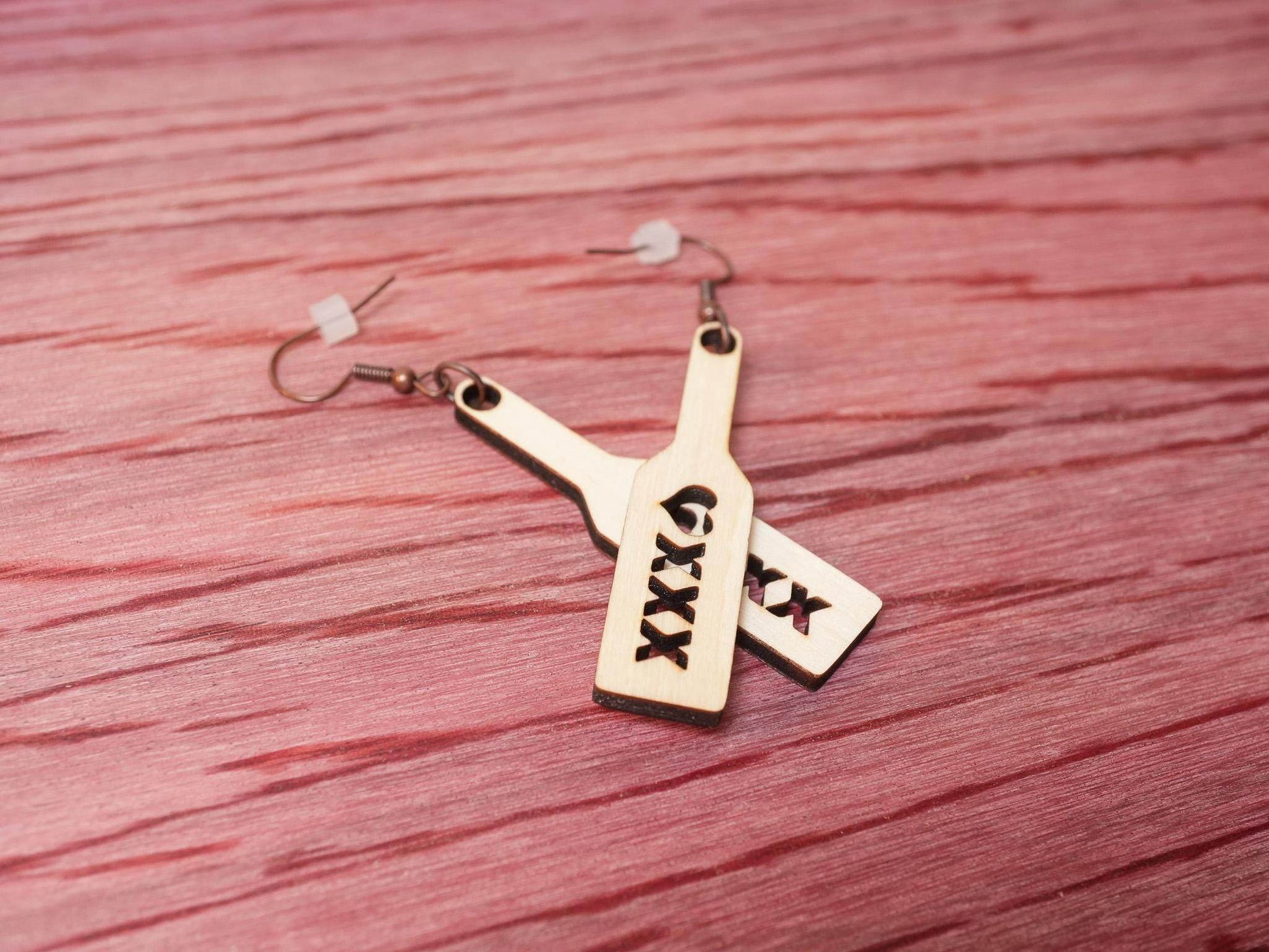 Bound by Dave wood spanking paddle earrings with triple X cutout design on a textured red surface