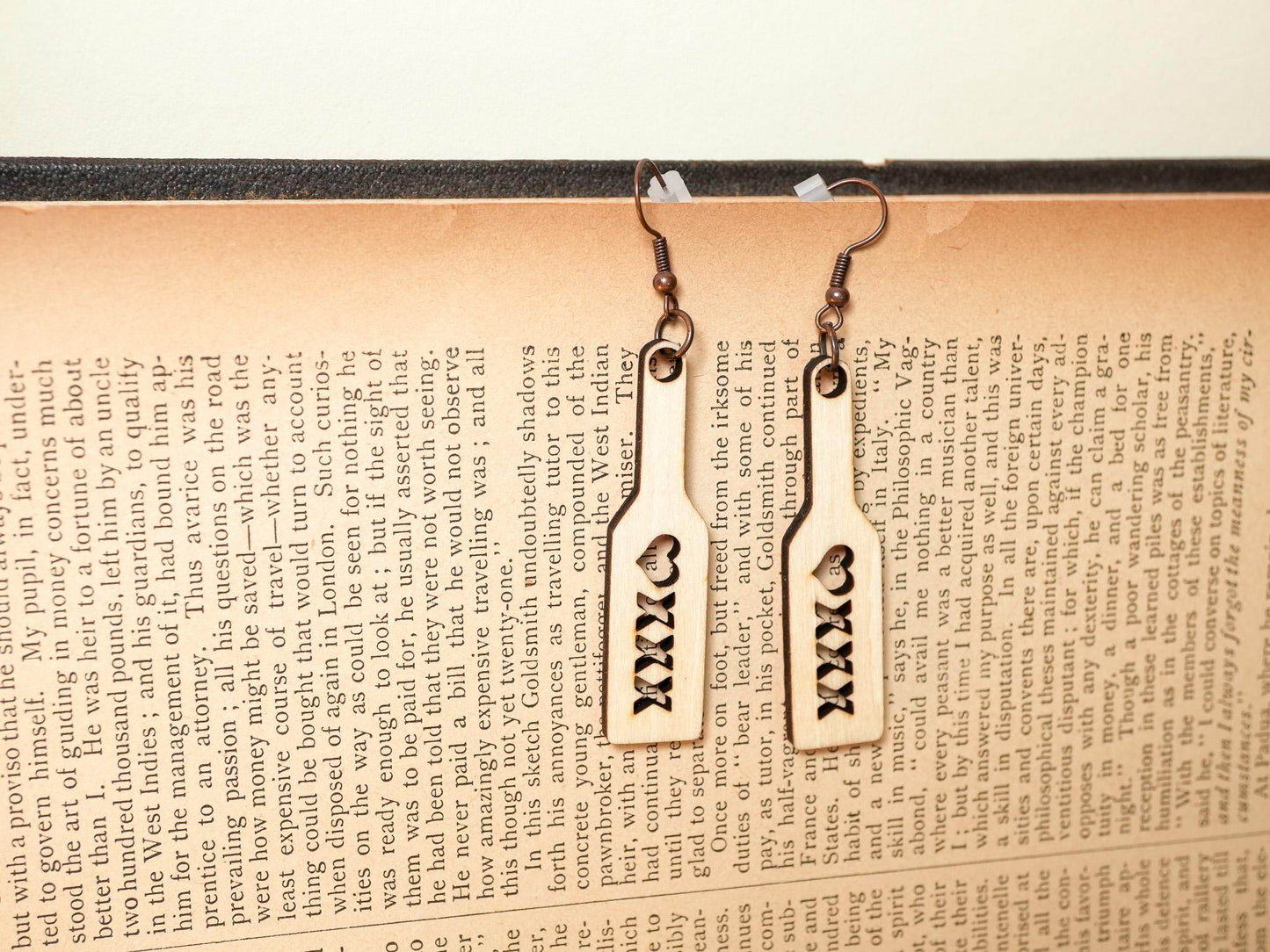 Bound by Dave wood spanking paddle earrings with heart cutout and XXX design hanging on a book page background