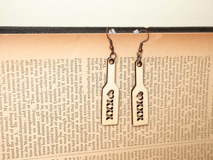 Bound by Dave wood spanking paddle earrings with heart cutout and XXX design hanging on a book page background