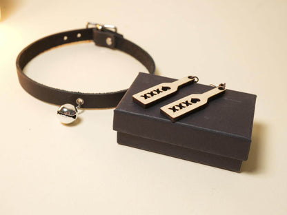 Wood spanking paddle earrings displayed on a black gift box beside a black leather choker with silver bell from Bound by Dave