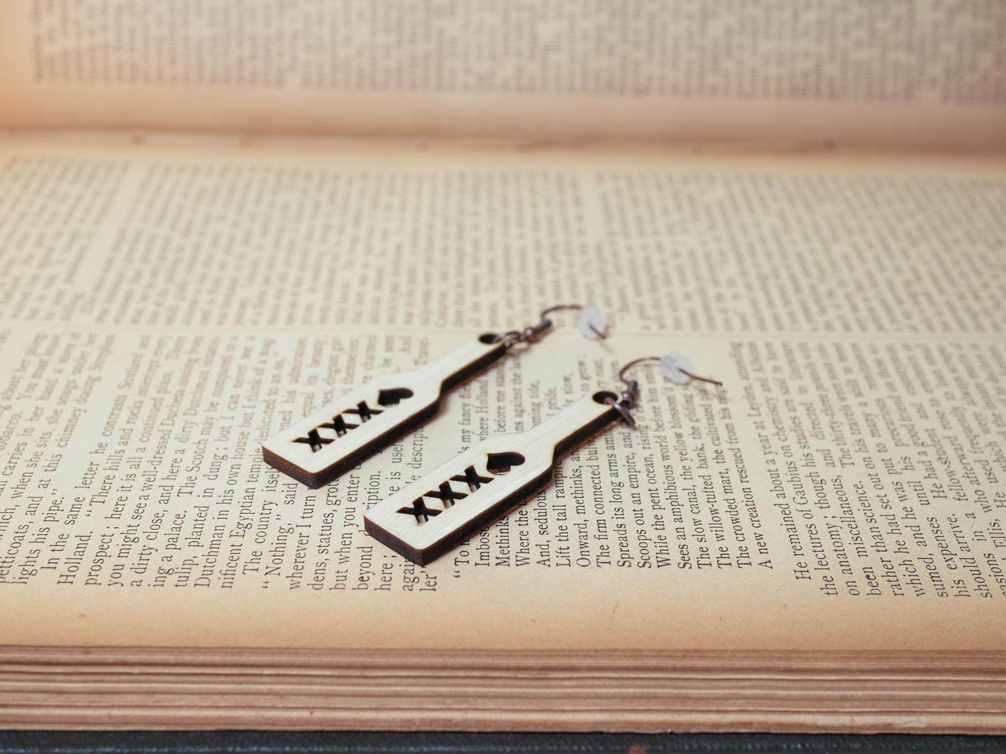 Bound by Dave wood spanking paddle earrings with black XXX and heart symbols on book background
